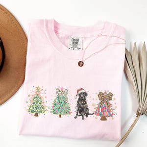 May include: Pink t-shirt with a Christmas-themed design featuring three decorated Christmas trees and a black Labrador Retriever wearing a Santa hat. The text "COMFORT COLORS" is visible on the tag.