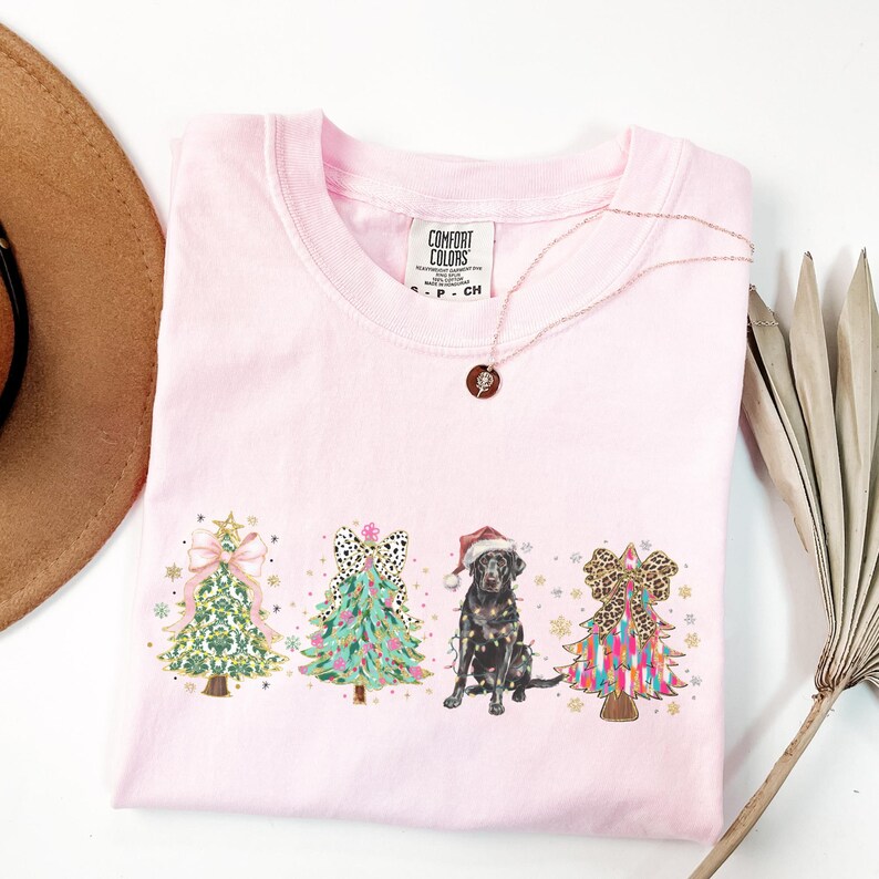 May include: Pink t-shirt with a Christmas-themed design featuring three decorated Christmas trees and a black Labrador Retriever wearing a Santa hat. The text "COMFORT COLORS" is visible on the tag.