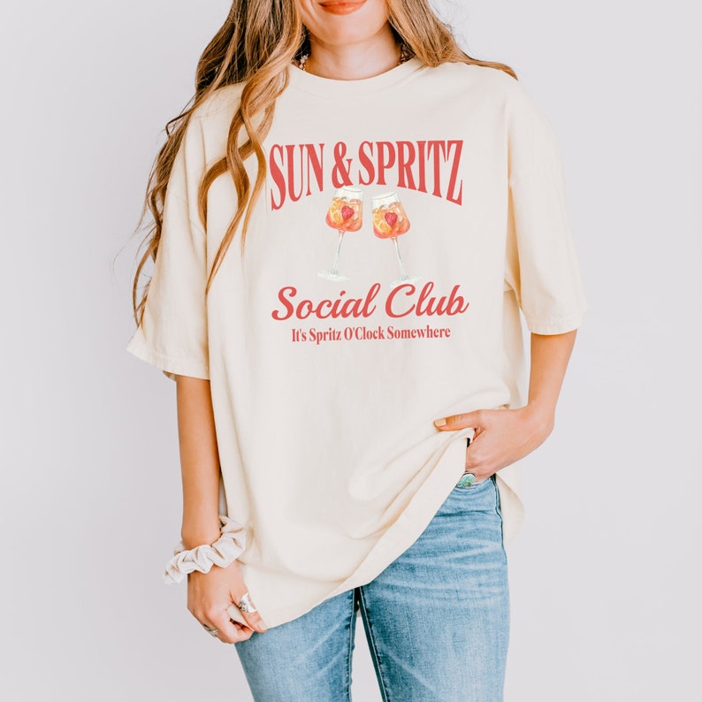 May include: A light yellow t-shirt with the text "Sun & Spritz Social Club It's Spritz O'Clock Somewhere" in red. The shirt features a graphic of two glasses of spritz.