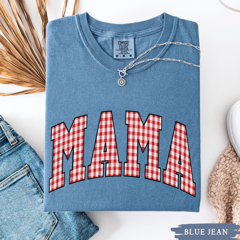 K&ouml;nnte beinhalten: Ein blaues Jeans-T-Shirt mit dem Wort "MAMA" in gro&szlig;en, rot-wei&szlig; karierten Buchstaben. Eine silberne Halskette mit Anh&auml;nger liegt &uuml;ber dem Shirt. Das Shirt ist gefaltet und liegt auf einer wei&szlig;en Oberfl&auml;che mit einer Jeans.