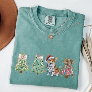 May include: A teal blue t-shirt with a festive Christmas design featuring three decorated Christmas trees and a corgi wearing a Santa hat and Christmas lights. The text "COMFORT COLORS" is visible on the tag.