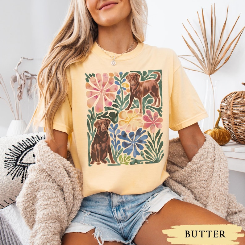 May include: Pale yellow t-shirt with a watercolour design of two brown dogs surrounded by colourful flowers and leaves. The shirt has a loose fit and short sleeves. The word "BUTTER" is printed at the bottom.