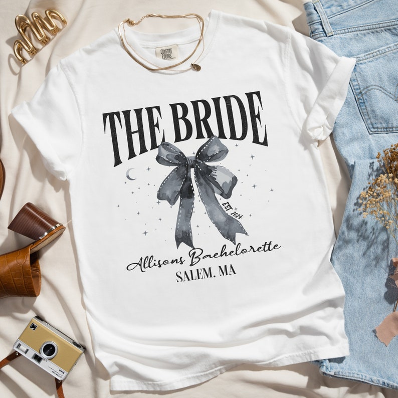 K&ouml;nnte beinhalten: Wei&szlig;es T-Shirt mit schwarzem Text, der "THE BRIDE" lautet, mit einer Aquarell-Schleife und Sternen. Der Text unter der Schleife lautet "Allison's Bachelorette SALEM, MA" und "Est. 2024".