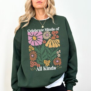 May include: Dark green sweatshirt with a floral design and the text "Celebrate Minds of All Kinds". The graphic features colorful flowers in shades of pink, orange, and yellow. The sweatshirt has a crew neck and long sleeves.
