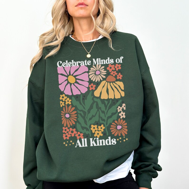 May include: Dark green sweatshirt with a floral design and the text "Celebrate Minds of All Kinds". The graphic features colorful flowers in shades of pink, orange, and yellow. The sweatshirt has a crew neck and long sleeves.