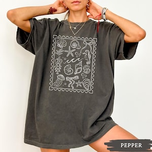 May include: A dark gray t-shirt featuring a white illustration of various sea creatures and elements, including seashells, starfish, and seahorses, within a rectangular frame. The shirt has short sleeves and a relaxed fit.