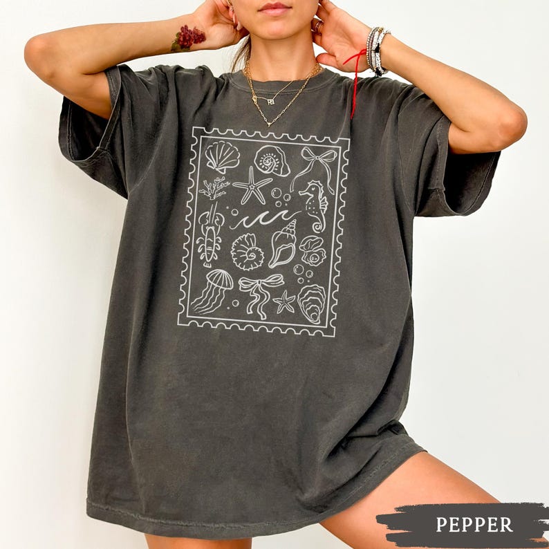 May include: A dark gray t-shirt featuring a white illustration of various sea creatures and elements, including seashells, starfish, and seahorses, within a rectangular frame. The shirt has short sleeves and a relaxed fit.