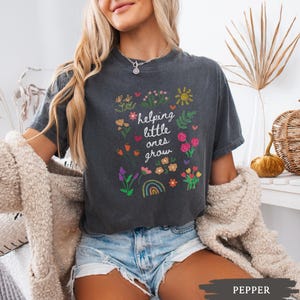 May include: A dark gray t-shirt with a white graphic that says "helping little ones grow" surrounded by colorful flowers, a rainbow, and a sun.