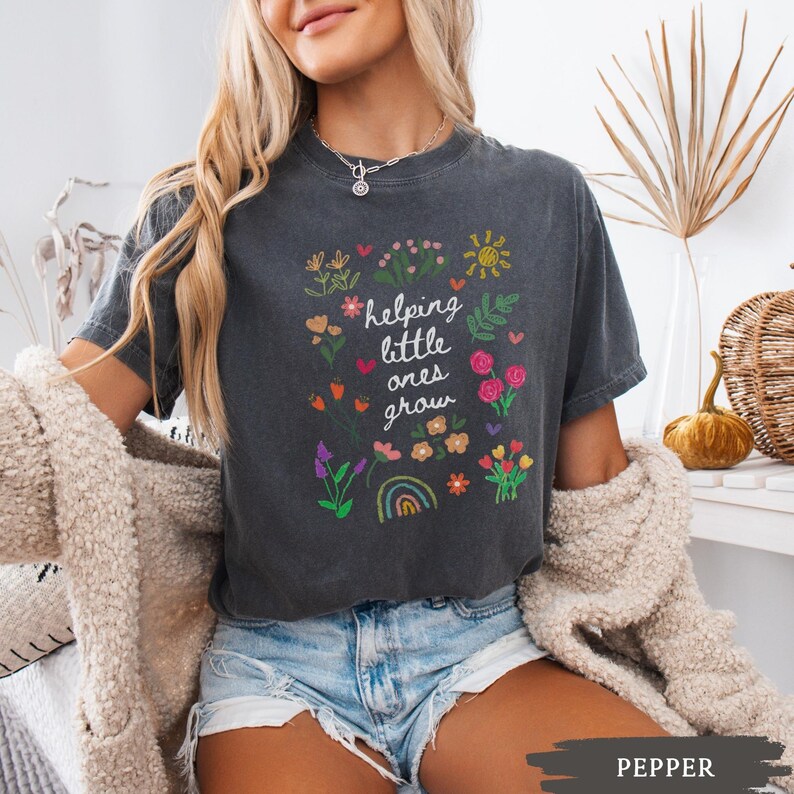 May include: A dark gray t-shirt with a white graphic that says "helping little ones grow" surrounded by colorful flowers, a rainbow, and a sun.