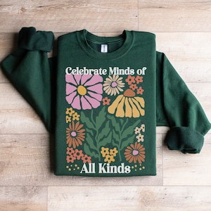 May include: A dark green sweatshirt with the text "Celebrate Minds of All Kinds" in white. The sweatshirt features a floral design with pink, yellow, orange, and white flowers. The sleeves are rolled up.