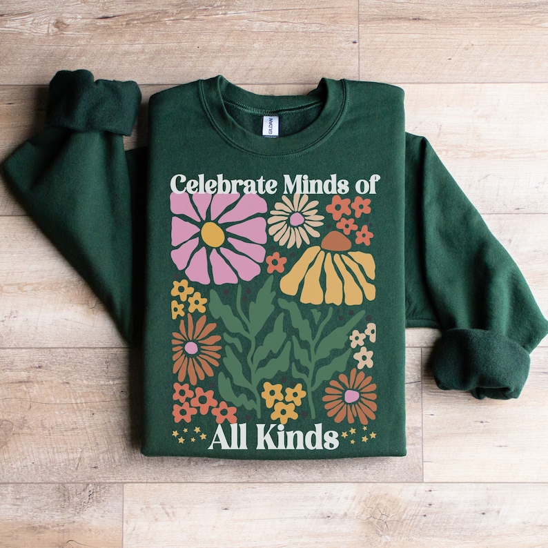 May include: A dark green sweatshirt with the text "Celebrate Minds of All Kinds" in white. The sweatshirt features a floral design with pink, yellow, orange, and white flowers. The sleeves are rolled up.