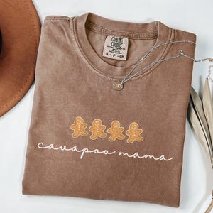 May include: A brown t-shirt with a white graphic of four gingerbread people and the text "cavapoo mama".