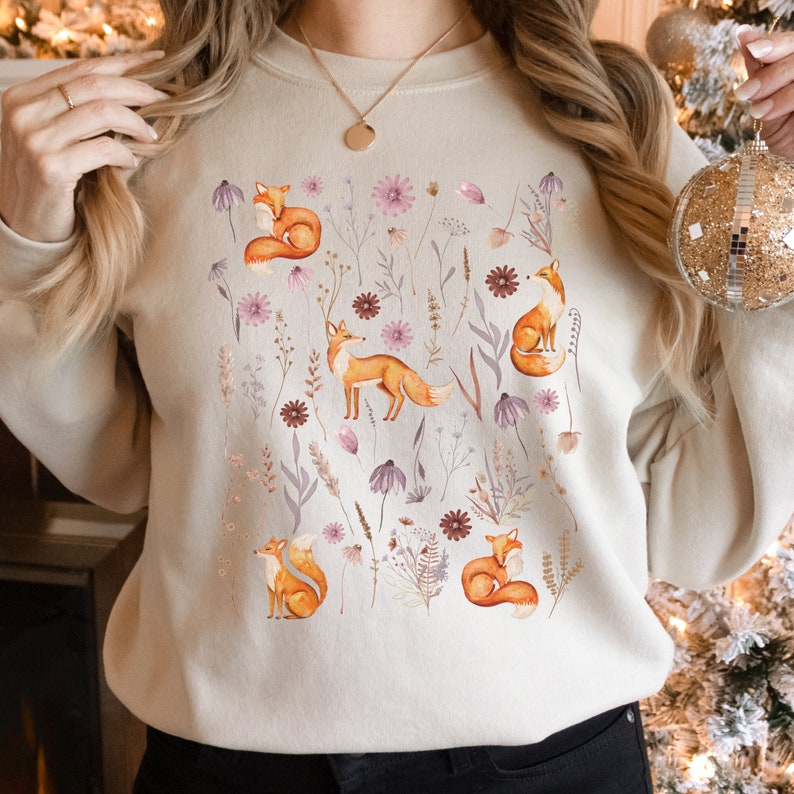 May include: A beige sweatshirt with a watercolor design of foxes and flowers. The foxes are orange and the flowers are pink, purple, and brown.