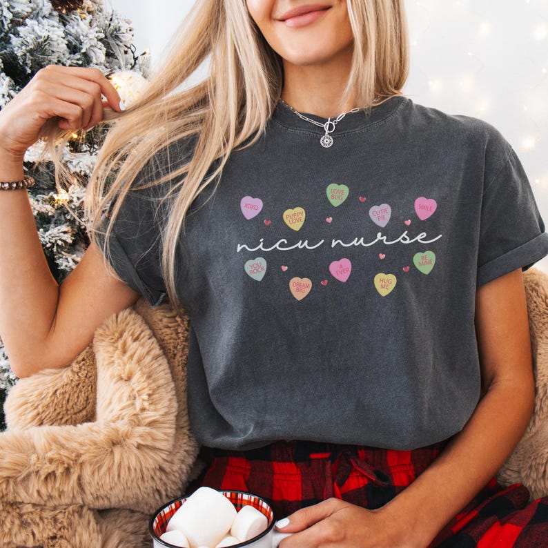 May include: A dark gray t-shirt with a white graphic of candy conversation hearts that say "XOXO", "Puppy Love", "Cutie Pie", "Be Mine", "You Rock", "I Love You", "Hug Me", "Pick Me", "Sweetheart", and "nicu nurse".