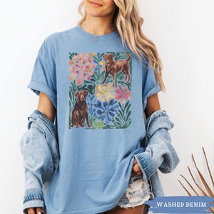 May include: Pale blue t-shirt with a watercolour design of two brown dogs surrounded by colourful flowers and leaves. The shirt has short sleeves and a loose fit. The words "WASHED DENIM" are printed at the bottom right.