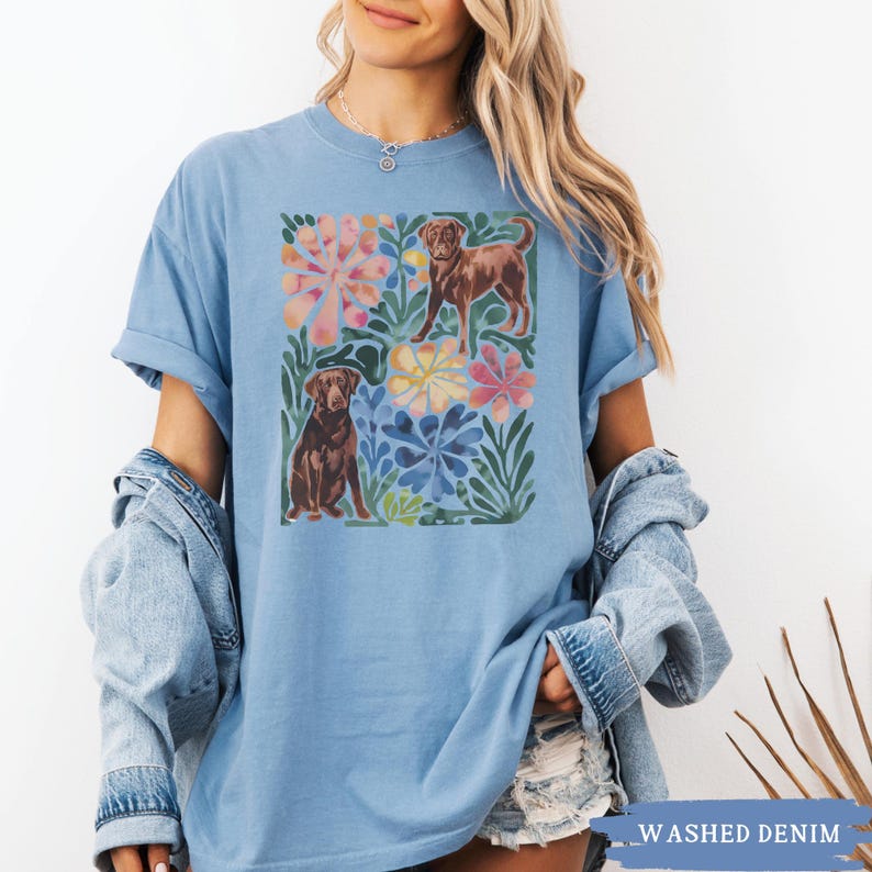 May include: Pale blue t-shirt with a watercolour design of two brown dogs surrounded by colourful flowers and leaves. The shirt has short sleeves and a loose fit. The words "WASHED DENIM" are printed at the bottom right.