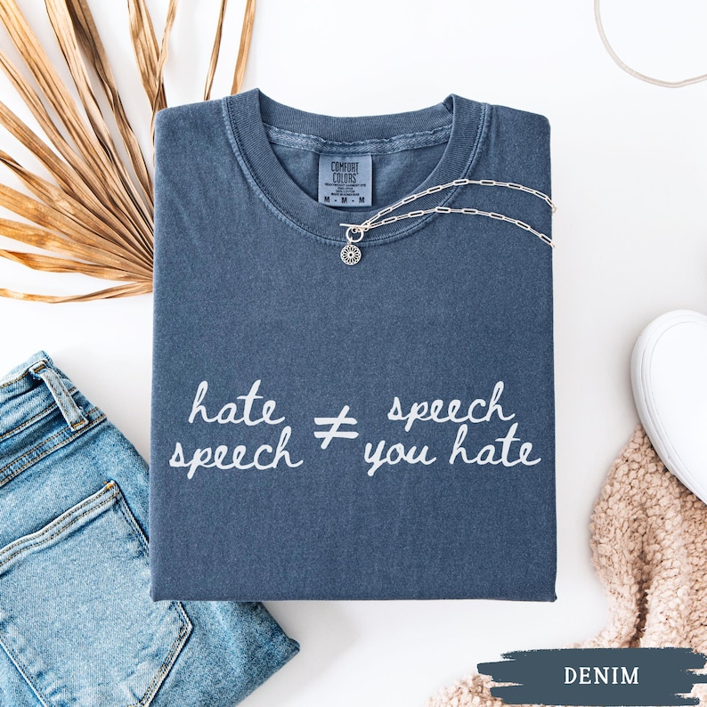 May include: A denim blue t-shirt with the text "hate speech &ne; you hate" in white script. A silver necklace with a small pendant rests on the shirt. The shirt is folded neatly, with a pair of jeans and a white shoe visible.