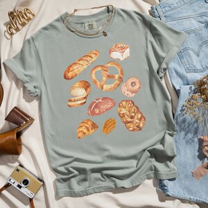 May include: A light green t-shirt with a watercolor design of various baked goods, including bread, pretzels, and pastries.