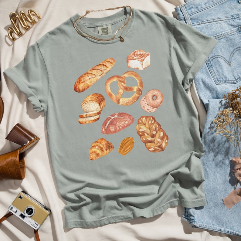 May include: A light green t-shirt with a watercolor design of various baked goods, including bread, pretzels, and pastries.