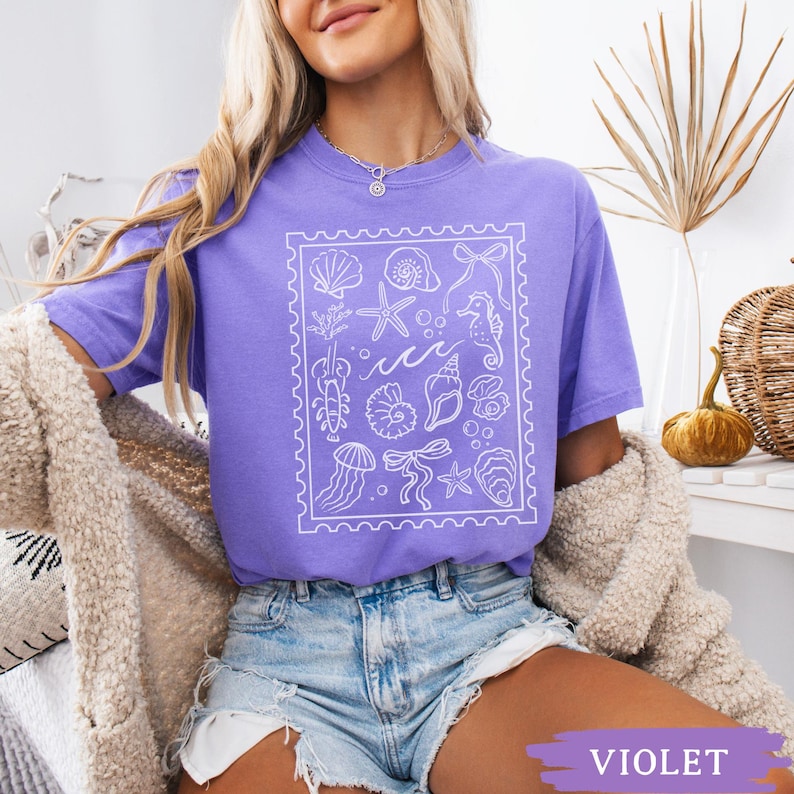 May include: Violet-colored t-shirt featuring a white graphic of various sea creatures and shells within a postage stamp-like border. The design includes starfish, seahorses, and jellyfish. The shirt is short-sleeved and made of a soft material.