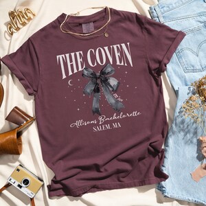 K&ouml;nnte beinhalten: Burgunderfarbenes T-Shirt mit einer wei&szlig;en Grafik, auf der "The Coven" mit einer schwarzen und grauen Schleife, einem Halbmond und Sternen steht. Der Text unter der Grafik lautet "Allison's Bachelorette Salem, MA Est. 2024".