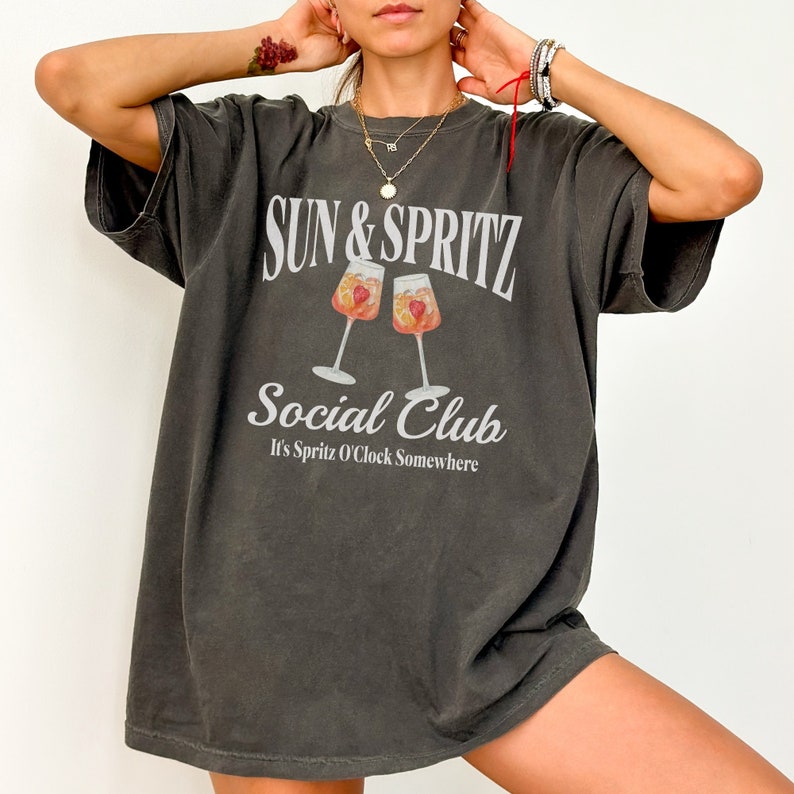 May include: A dark gray oversized t-shirt with the text "SUN & SPRITZ Social Club It's Spritz O'Clock Somewhere" and a graphic of two glasses of spritz.