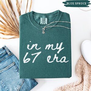 May include: A folded blue spruce Comfort Colors t-shirt with the text "in my 67 era" in white script. A silver necklace with a sunburst pendant rests on the shirt. A pair of blue jeans, a white shoe, and a tan scarf are also in the image.