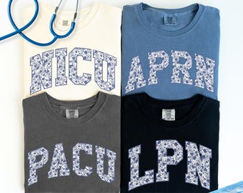 May include: Four t-shirts with different medical abbreviations printed on them. The abbreviations are NICU, APRN, PACU, and LPN. The shirts are all different colors, including white, blue, gray, and black. The shirts are all folded and laid out on a white surface. A stethoscope and a blue face mask are also visible in the image.