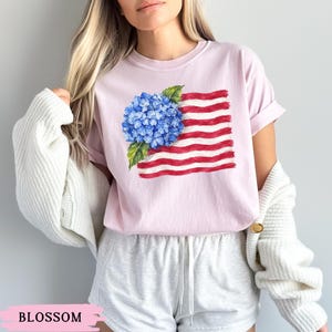 Op de afbeelding: Een lichtroze T-shirt met korte mouwen en een aquarel-stijl afbeelding van een blauwe hortensia bovenop horizontale, golvende rode en witte strepen. Het shirt heeft een ronde hals en een ontspannen pasvorm.