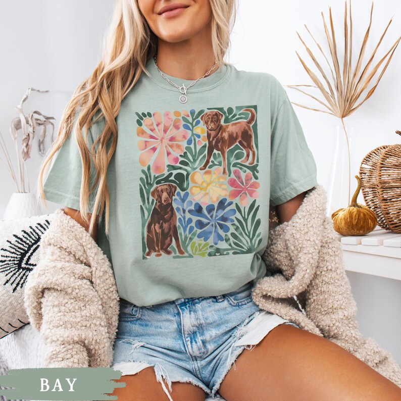 May include: A pale green t-shirt with a watercolour design of two brown dogs amidst colourful flowers and green leaves. The word "BAY" is printed at the bottom. The shirt is made of a soft material.
