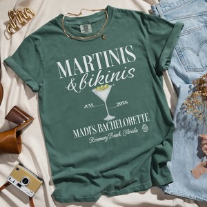 May include: Green t-shirt with white text that reads "Martini's & bikinis" with a martini glass illustration and the text "June 2026 Madi's Bachelorette Rosemary Beach, Florida".