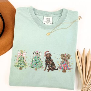 May include: A light green t-shirt featuring a Christmas tree design with a chocolate Labrador dog and three decorated Christmas trees. The trees are decorated with bows, lights, and baubles. The shirt has the text "Comfort Colors" on the tag.
