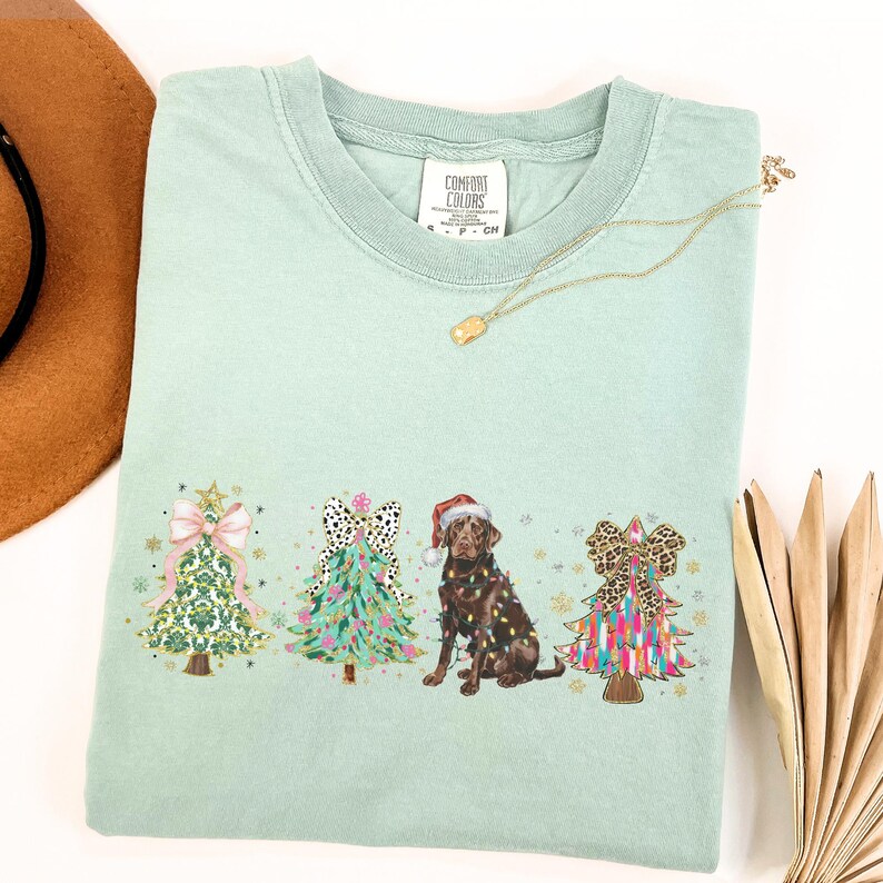 May include: A light green t-shirt featuring a Christmas tree design with a chocolate Labrador dog and three decorated Christmas trees. The trees are decorated with bows, lights, and baubles. The shirt has the text "Comfort Colors" on the tag.