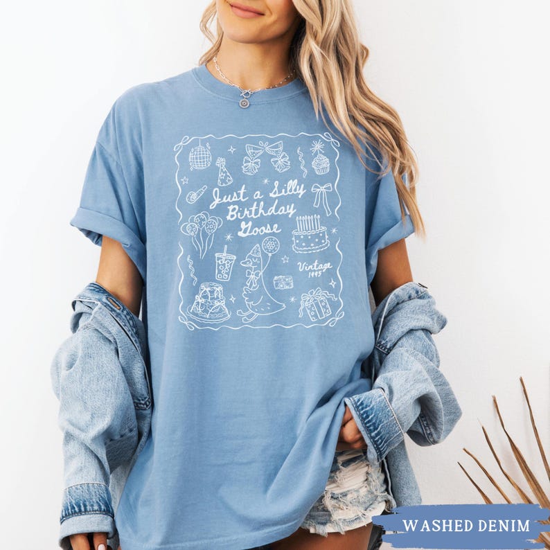 May include: A light blue t-shirt with a white graphic that says "Just a Silly Birthday Goose" with birthday-themed illustrations. The shirt is a washed denim color and has rolled-up sleeves.