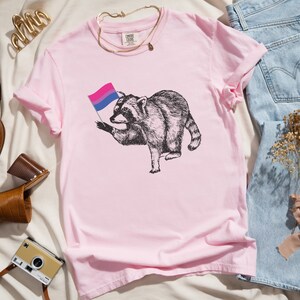 May include: Pink t-shirt with a black and white illustration of a raccoon holding a bisexual pride flag.