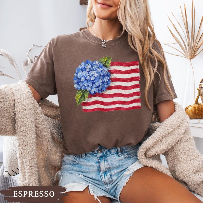 Op de afbeelding: Een bruin t-shirt met korte mouwen en een grafische print van een blauwe hortensia-bloemtros bovenop een gestileerde Amerikaanse vlag met rode en witte strepen. De tekst ESPRESSO staat in het wit onderaan de afbeelding gedrukt.