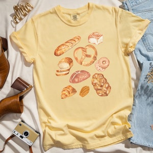 May include: A yellow t-shirt with a watercolor illustration of various breads, including a baguette, pretzel, and rolls.