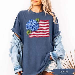 Op de afbeelding: Een donkerblauw T-shirt met korte mouwen en een grafische print van een blauwe hortensiabloem geplaatst over een gestileerde Amerikaanse vlag met rode en witte horizontale strepen. Het T-shirt heeft een ronde hals en een losse pasvorm.