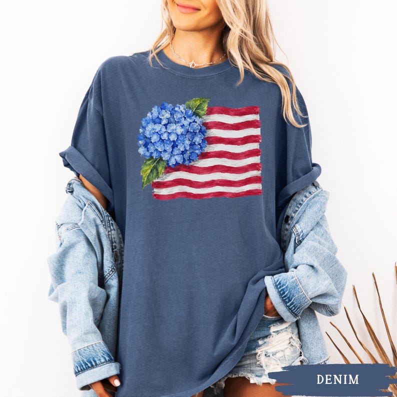 Op de afbeelding: Een donkerblauw T-shirt met korte mouwen en een grafische print van een blauwe hortensiabloem geplaatst over een gestileerde Amerikaanse vlag met rode en witte horizontale strepen. Het T-shirt heeft een ronde hals en een losse pasvorm.