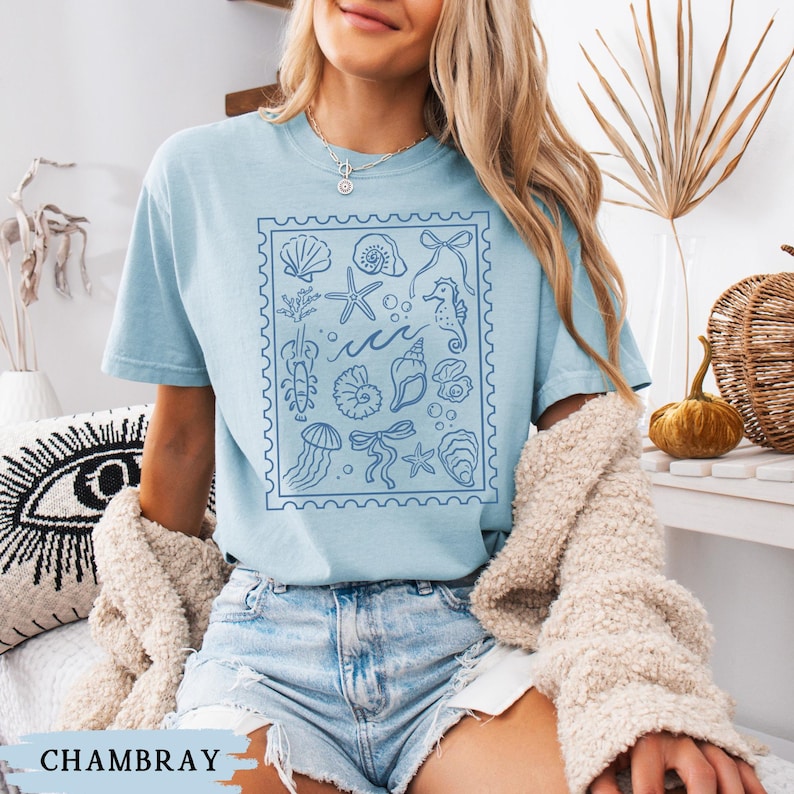 May include: Light blue t-shirt with a blue stamp-like design featuring various sea creatures and shells. The word "Chambray" is printed at the bottom. The shirt is a casual, short-sleeved style.