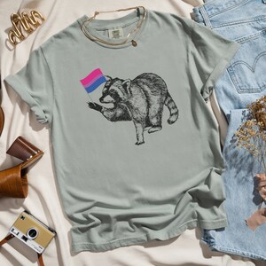 May include: A light blue t-shirt with a graphic of a raccoon holding a bisexual pride flag. The flag is pink, blue, and white.