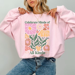 May include: A light pink sweatshirt with a floral design and the text "Celebrate Minds of All Kinds" in green. The floral design includes pink, yellow, orange, and brown flowers with green leaves.