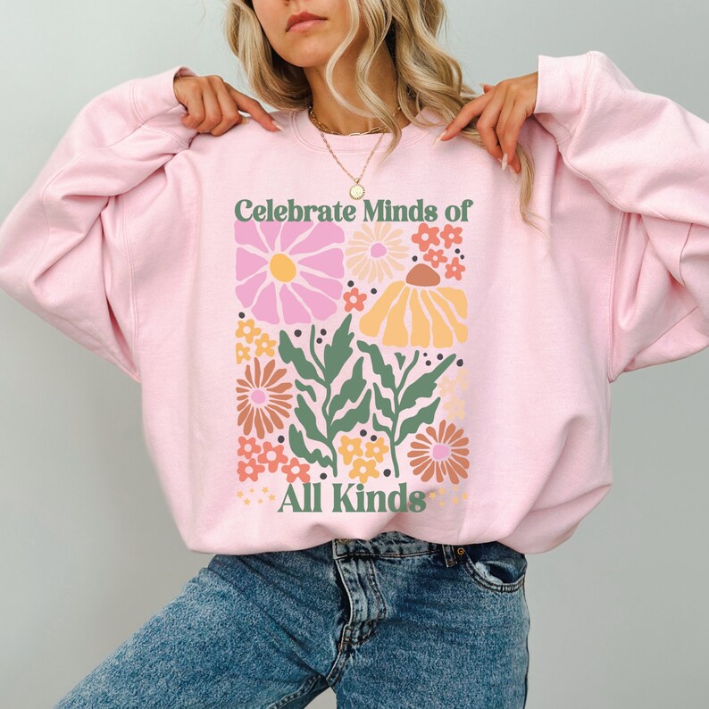 May include: A light pink sweatshirt with a floral design and the text "Celebrate Minds of All Kinds" in green. The floral design includes pink, yellow, orange, and brown flowers with green leaves.