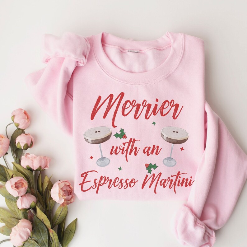 May include: Pink sweatshirt with the text "Merrier with an Espresso Martini" in red lettering. Two martini glasses with coffee and holly are on either side of the text.