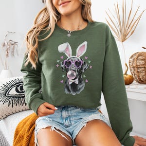 May include: A green sweatshirt featuring a black dog wearing pink heart-shaped sunglasses and bunny ears. The dog is blowing a pink bubble. The sweatshirt has a small bow tie on the dog's neck and pink hearts and crosses around the dog.