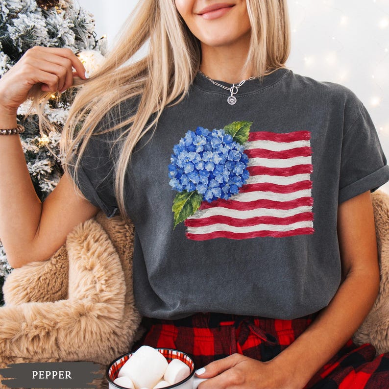 Op de afbeelding: Een antracietgrijs T-shirt met korte mouwen en een grafische print van een blauwe hortensiabloem over een gestileerde Amerikaanse vlag met rode en witte horizontale strepen, voorzien van een ronde hals en een relaxte pasvorm.