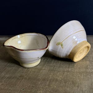 May include: Two small ceramic bowls with a cream-colored glaze and a light yellow base. One bowl is positioned upright, showing a dark brown rim, while the other is tilted to reveal a small green leaf motif painted on the exterior side.