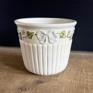 May include: A white ceramic planter featuring a raised relief pattern around the upper rim. The design includes sculpted light blue bows, green ivy leaves, and small red berries. The lower section of the planter has vertical fluted ridges.