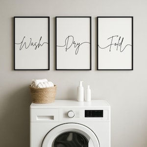Può includere: Tre stampe incorniciate con le parole "Wash", "Dry" e "Fold" in corsivo nero. Sotto, una lavatrice bianca con un quadrante rotondo e un cestino intrecciato con asciugamani piegati. Due bottiglie bianche sono sulla macchina.