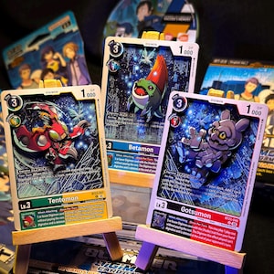 May include: Three Digimon trading cards are displayed on miniature wooden easels. The cards feature colorful illustrations of Digimon characters, with text and numerical values. The cards are set against a dark background, with additional Digimon card packs visible.